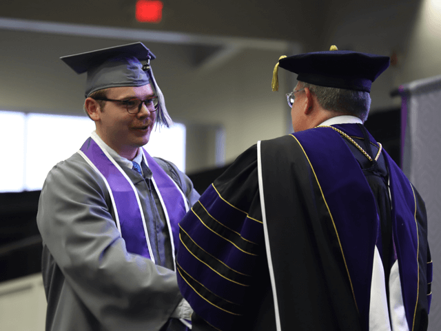 male student in gray graduation gown shakes president's hand and receives diploma on graduation stage