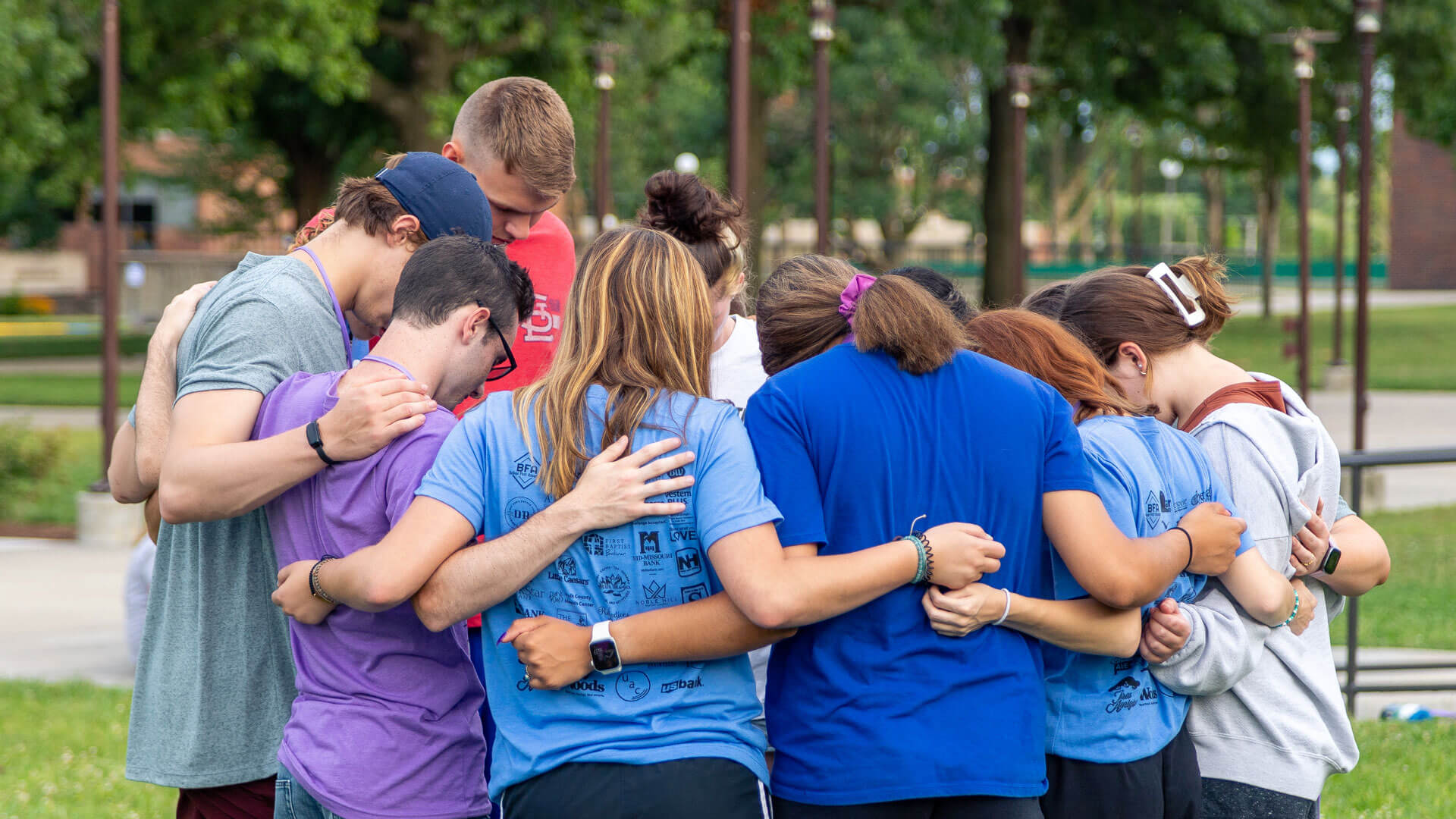 students gather in huddle with arms around each other and heads bowed in prayer