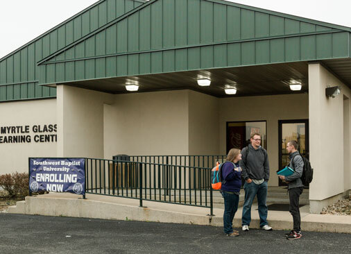 three students stand outside campus building talking to each other
