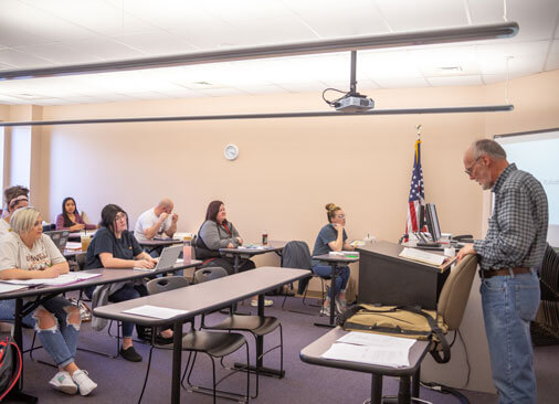 teacher stands in front of rows of students sitting at tables