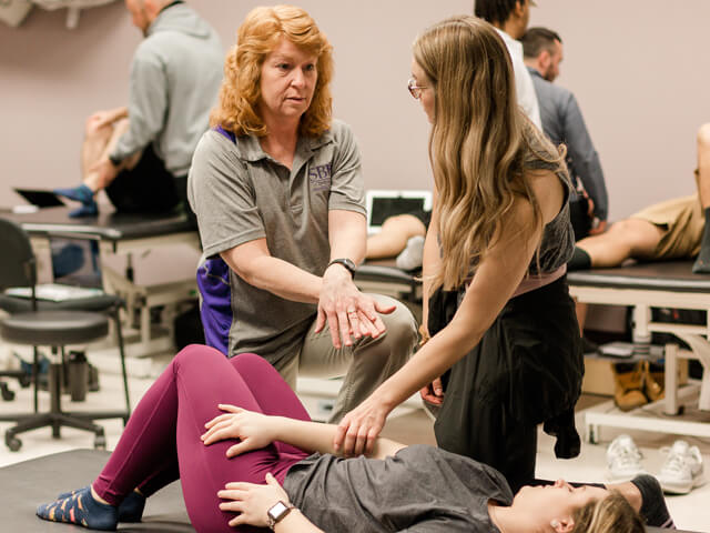 Physical therapy professor demonstrates an exercise on a student laying on lab table while explaining to another student looking on