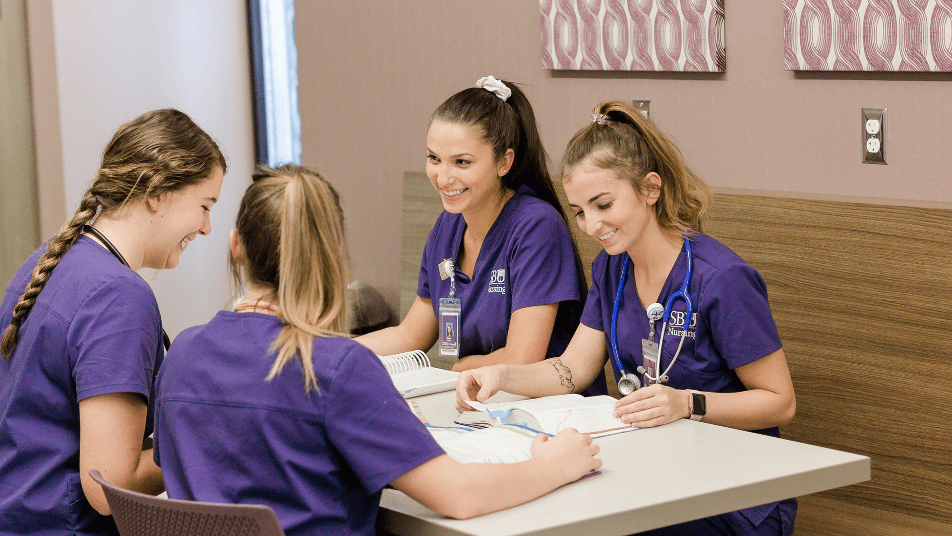 four girls wearing purple scrubs smile and laugh as they sit at a table studying together