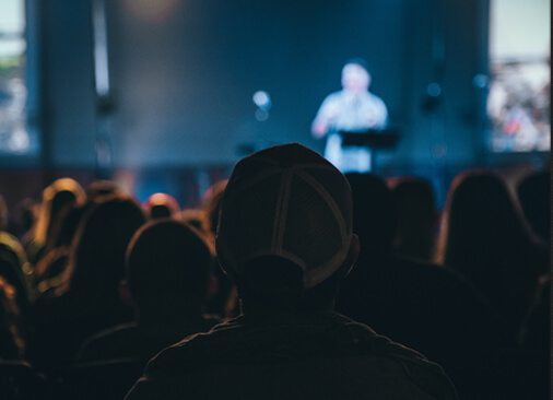 blurred image from back of people sitting in conference with speaker on stage